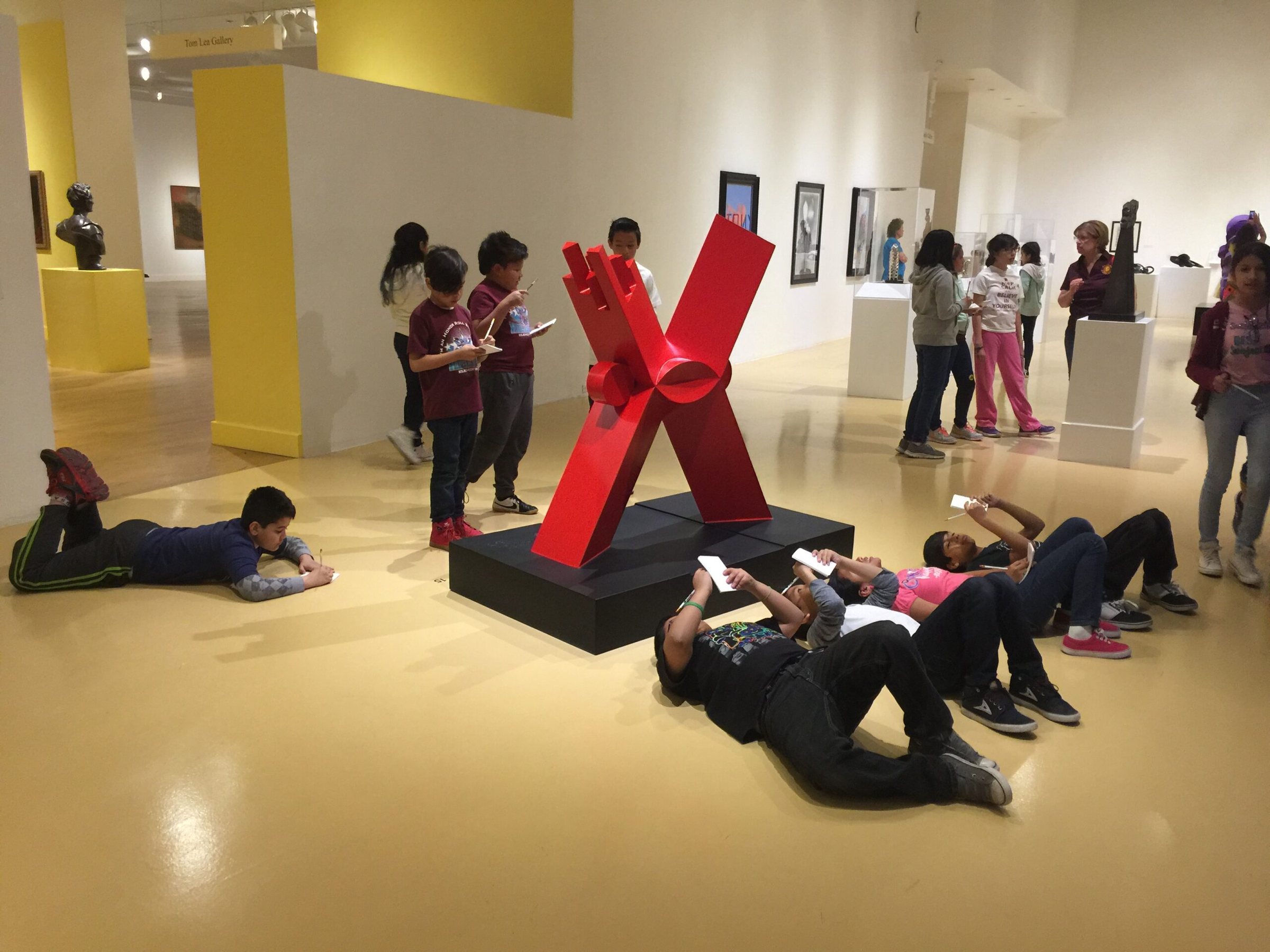 Elementary students sketching in notebooks around a large red steel sculpture in a gallery at the El Paso Museum of Art, several lying on the floor to draw.