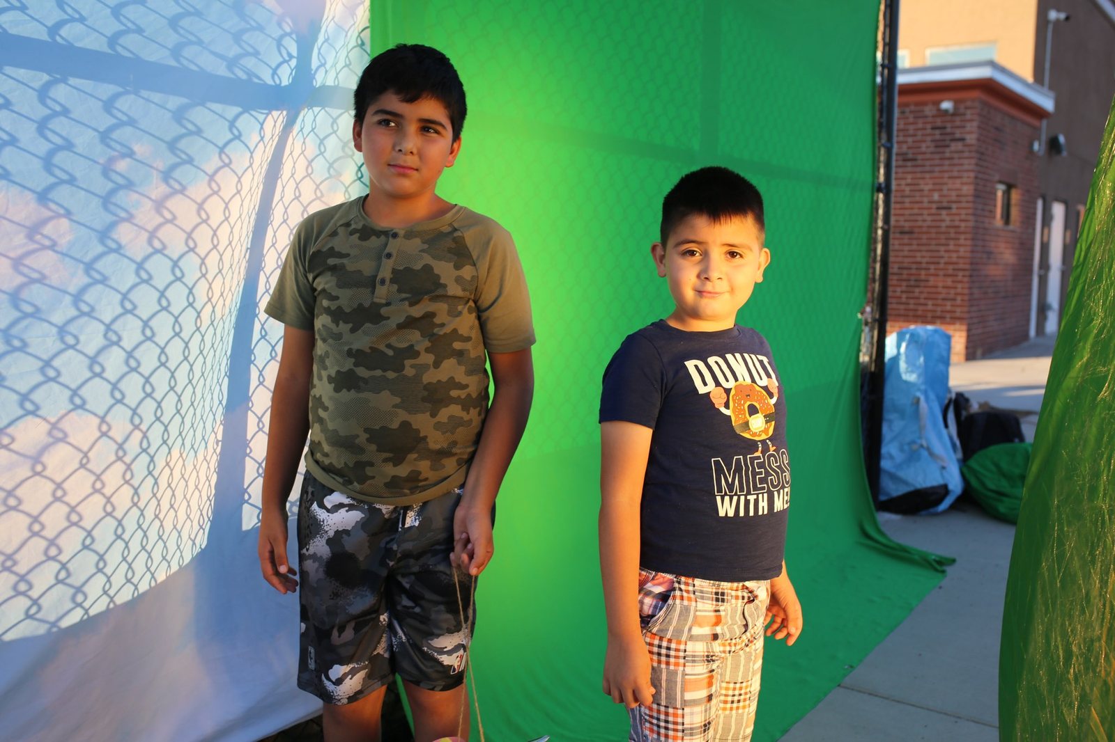 Two boys standing in front of a green screen set up outdoors at an El Paso public housing complex during a media workshop, in late afternoon light.